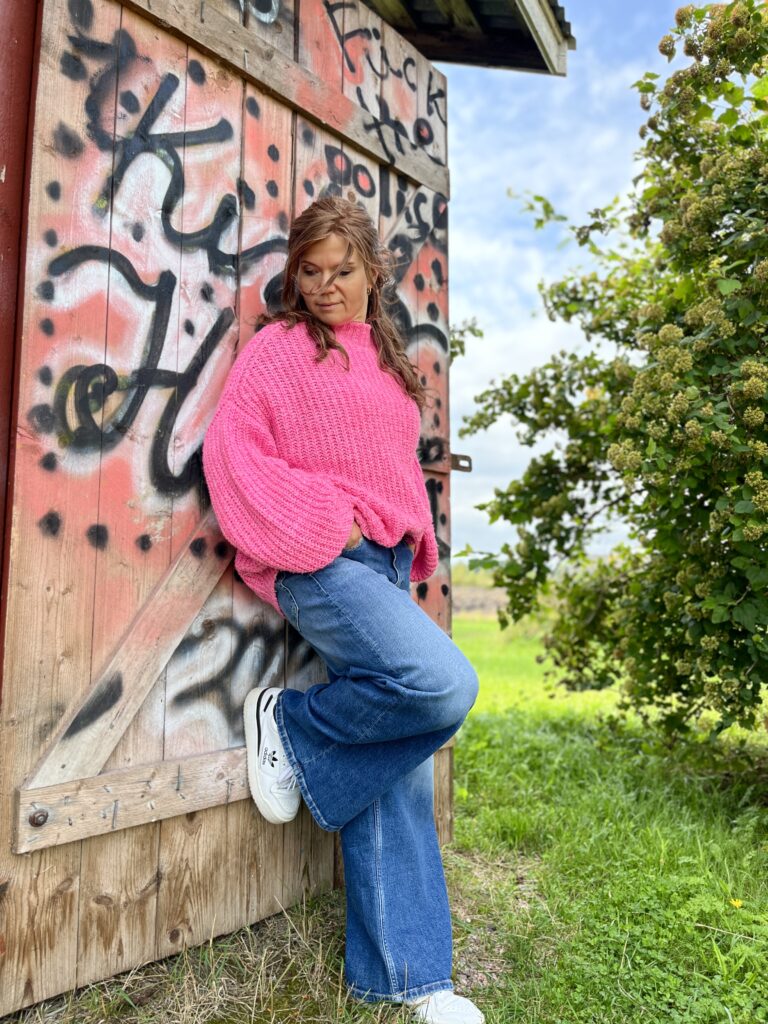 Henkilö linkissä villapaidassa nojaamassa graffitti seinään vihreässä  ympäristössä